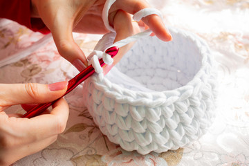 Young woman while crocheting on a patterned tablecloth, close up. Stay at home leisure activity idea. Basket made of white T-shirt yarn, with red crochet needle. Pale pink nails. - obrazy, fototapety, plakaty