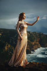 Beautiful red-haired girl in a gold dress and crown stands on the shore of the ocean