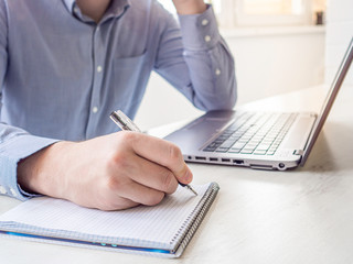 Close up mans hand writing notes with pen. Man speaking mobile phone, working on laptop at home. Concept of business, working from home, quarantine, isolation, coronavirus.