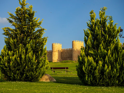 Parte de las Murallas de Avila entre dos grandes setos verdes y un banco de madera en medio con cielo azul