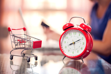 Woman doing online shopping at home. Laptop, alarm clock and shopping trolley on table.
