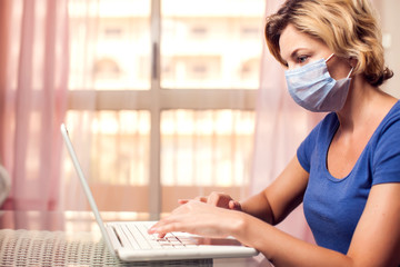 Woman with medical face mask working with laptop at home. Self-isolation while pandemic of coronavirus