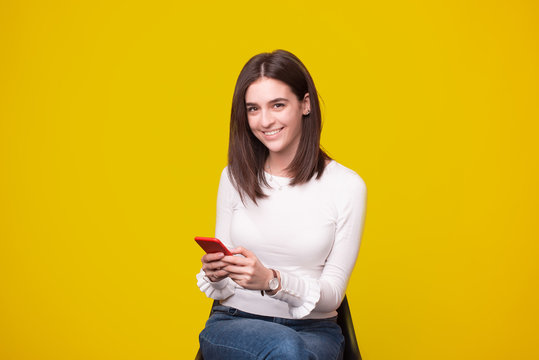 Photo of sitting girl looks at the camera while typing or surfing her phone on yellow background.