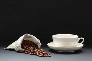 fresh coffee beans sprinkled from a bag on a black background next to a white cup