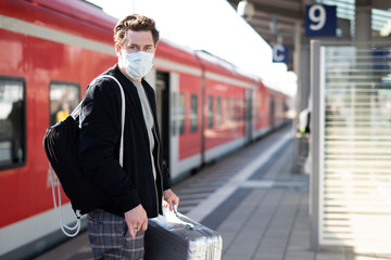 Mann mit Koffer auf dem Bahnsteig, er schützt sich mit Mundschutz und Handschuhen vor einer...