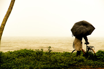 old man on his bike looks at the horizon and the sea with his umbrella broken by the wind, on the coast, South India