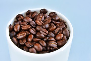 Roasted coffee beans in porcelain mug with selective focus on neutral background. Arabica coffee seeds in the coffee cup