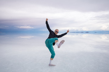 Sun Burnt Caucasian Bold Man with Sunglasses in Black Shirt and Green Pants Poses in Flooded Uyuni Salt Flats at Sunset