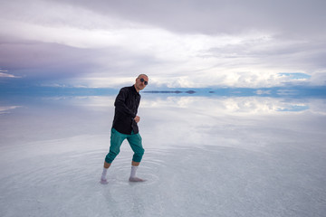 Sun Burnt Caucasian Bold Man with Sunglasses in Black Shirt and Green Pants is Dancing in Flooded Uyuni Salt Flats at Sunset