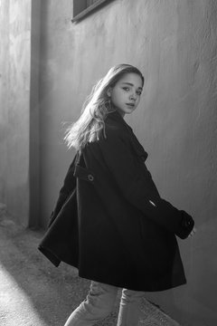 Young beautiful girl with long hair in black coat on sunny day turns around against the wall. Street style portrait photosession. Black and white image