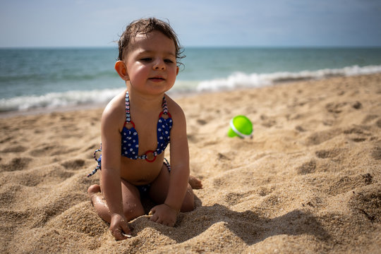 little cute toddler tanned Caucasian girl in a blue swimsuit sitting on a sandy beach against the background of sea waves