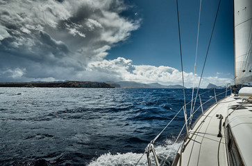 The view of the sea and mountains from the sailboat, edge of a board of the boat, slings and ropes, splashes from under the boat, sunny weather, dramatic sky