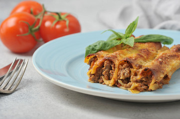 Cannelloni with minced meat on a blue plate. Cutlery and ripe tomatoes complete the composition. Light background. Close-up.