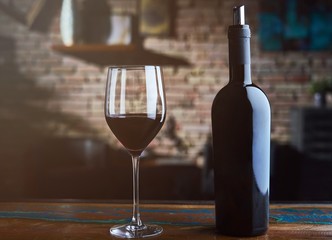 Glass of red wine and wine bottle on colorful wooden table at home, brick wall in the background.