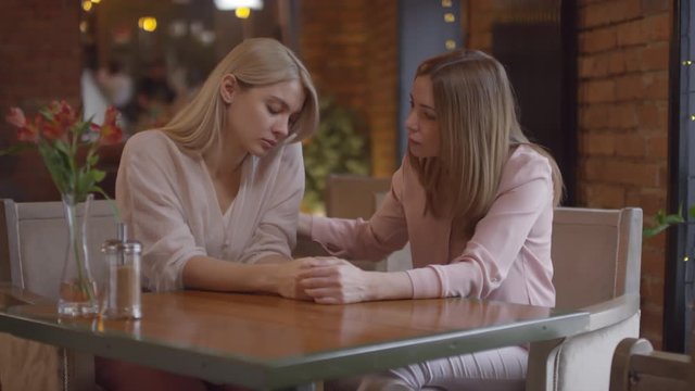 Handheld shot of supportive mother holding hands with young blond daughter and giving her words of encouragement while they are sitting at table in cafe