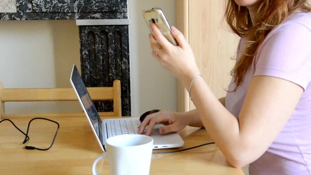 Serious young woman freelancer working on freelance from home typing email on laptop, girl using computer for study online uses a phone, drinks coffee or tea, female user busy on distance internet job