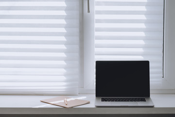Fototapeta premium Laptop with a blank screen and a notebook on the window on a sunny day. Workplace home