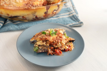 Moussaka served with parsley garnish on a blue gray plate on a white table, traditional Greek casserole of eggplants, potatoes, minced meat and tomatoes, copy space