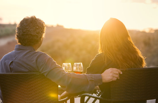 Man And Woman Sitting With Wine Glasses Overlooking Vinyard At Sunset