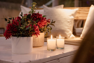 Decoracion de sala de estar con sillas de mimbre, mubles de madera, velas, flores rojas y almohadones blancos.