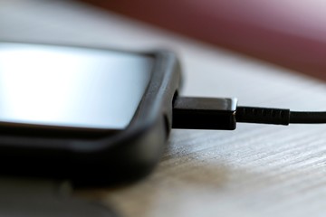 A close up portrait of a smartphone in a sturdy case charging on a wooden table with a USB cable in its port. The phone had to charge because its battery died.