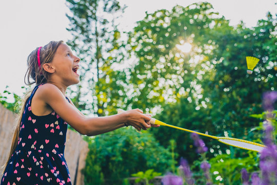 Young Caucasian Girl Or Kid Is Playing Badminton Game In A Lush Green Garden. Kid Laughing In A Garden While Holding A Racket