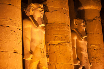 Night view of dramatically lit stone statues of ancient Egyptian pharaoh Ramses II standing between colossal columns in Luxor, Egypt
