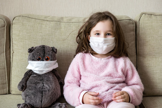 COVID-19 coronavirus and quarantine concept, kid in medical mask with toy on couch at home. Little girl in room during corona virus pandemic.