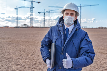 civil engineer in a medical mask and white helmet on the background of construction, with a raised...