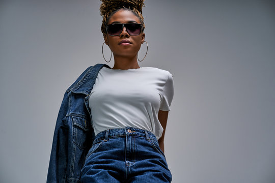 Confident lady in sunglasses in white t-shirt in jeans with denim jacket posing on camera with chest forward and holds hands behind her back. Fashion concept