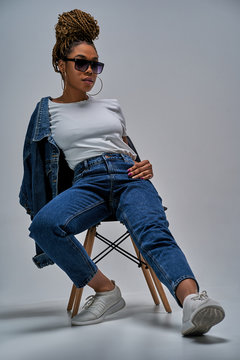 Confident lady in sunglasses in white t-shirt in jeans with denim jacket posing on camera sitting on chair with leg forward. Fashion concept