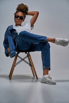 Confident lady in sunglasses in white t-shirt in jeans with denim jacket posing on camera sitting on chair with leg lies on the leg. Fashion concept
