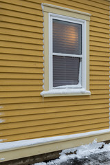 The exterior of a yellow coloured wall of narrow clapboard. In the center of the building, there's a double-hung window that is closed and has snow on the ledge and light at the top of the window. 