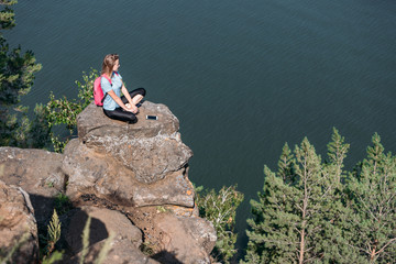 A young blonde girl with a backpack and a smartphone sits on a rocky ledge in the mountains against the backdrop of a sea landscape.