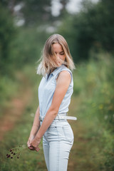 Fototapeta premium A young blonde girl stands on a forest road with flowers in her hands and turns her head. Hair fluttering in the wind.