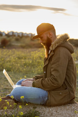 Modern young man sitting in the field working at sunset with his latop