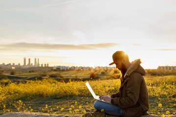 Young modern traveller working with his laptop in the countryside at sunset with the city skyscrapers in the background