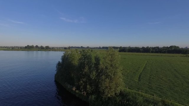 Smooth aerial drone shot of some trees standing  on the waterline of a lake in the grasslands of the Netherlands