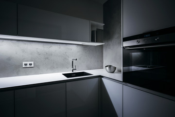 Modern white kitchen in minimalist design, in night, with light strip turned on, modern oven, granite sink, wall-sockets and premium materials such as glass, concrete, aluminum and stainless steel. - obrazy, fototapety, plakaty