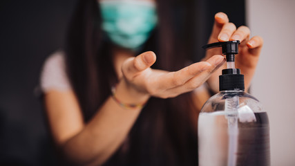 Young asian woman with long hair wearing medical mask and sanitizing her hands at home. Concept of coronavirus. Horizontal image, selectvie focus on hands and bottle of hand sanitizer gel.