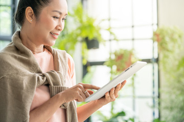 Beautiful Asian senior woman enjoy using digital tablet for online shopping or reading social media news in greenhouse garden at home. Elderly leisure activity lifestyle and healthcare concept.