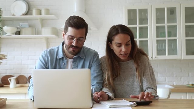Pleasant focused young mixed race family spouses calculating payments, monthly expenses, using computer banking app. Married couple managing budget, counting bills, doing paperwork together at home.