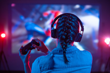 Girl in headphones plays a video game on a screen projector or TV in the dark room. Gamer with a joystick. Online games with friends, competitions. Fun entertainment. Teenagers play arcade. Back view - obrazy, fototapety, plakaty