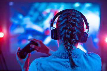 Girl in headphones plays a video game on a big screen projector or TV in a dark room. Gamer with a...