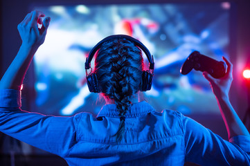 Young girl in headphones plays a video game on the TV in the evening. Gamer with a joystick. Online gaming with friends, competitions, win. Fun entertainment. Teens play puzzle games. Back view - obrazy, fototapety, plakaty