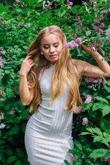 Portrait of a charming blond woman wearing beautiful white dress standing next to lilac bush.