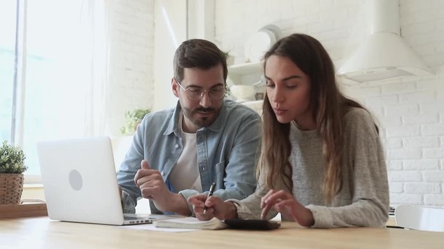 Focused young wife calculating mortgage payments, writing notes, while handsome husband in eyeglasses using online banking application. Concentrated couple managing monthly family budget together.