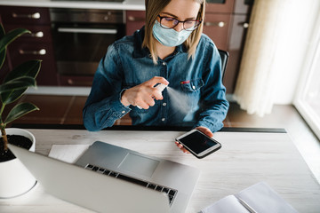 Coronavirus. Sanitizer. Business woman working in home, wearing protective mask in quarantine. Cleaning hands mobile phone to eliminate germs antibacterial spray. Stay at home. Prevent Covid-19 virus.