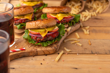 Juicy Burger on a wooden table with a drink. Homemade cheeseburger on the cutting board with beef, cheese, onions, tomatoes, cucumbers, sauce, french fries and sweet soda. Concept of street food
