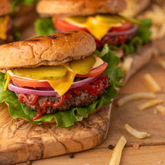 Hot cheeseburger on a wooden cutting board. Burger with beef, cheese, vegetables, crispy buns and salt fries on a rustic table. Fast food concept. Front view